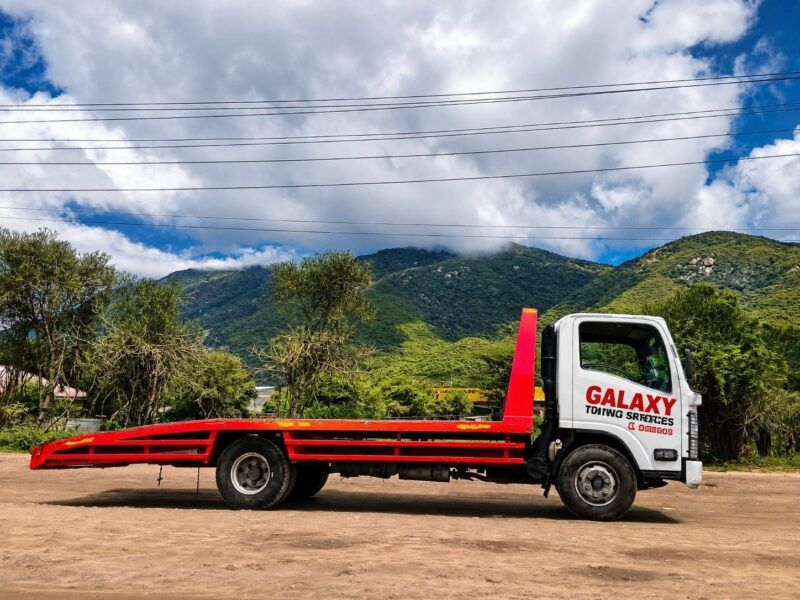 A flatbed tow truck labeled GALAXY TOWING SERVICES, specializing in flatbed towing and breakdown services in Runda, is parked on a dirt road with green trees and mountains in the background under a partly cloudy sky.
