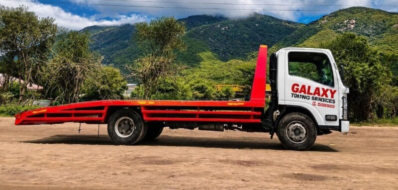 A flatbed tow truck labeled GALAXY TOWING SERVICES, specializing in flatbed towing and breakdown services in Runda, is parked on a dirt road with green trees and mountains in the background under a partly cloudy sky.